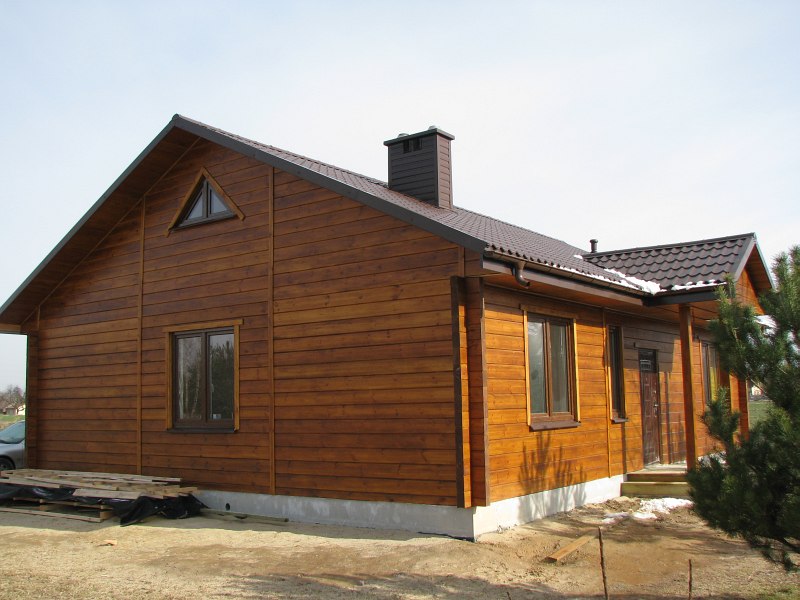 Gotowy dom z drewna o powierzchni 96 m² z brązowym dachem i kominem, widok z boku na tle zimowego krajobrazu z resztkami śniegu.